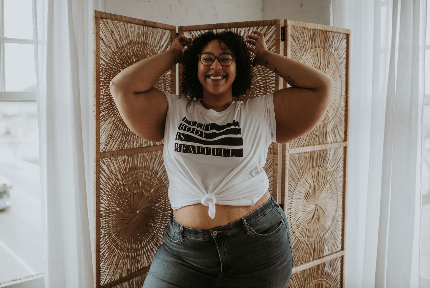 A joyful figure poses against a sunlit wall with macrame decor, wearing denim and a white crop top.