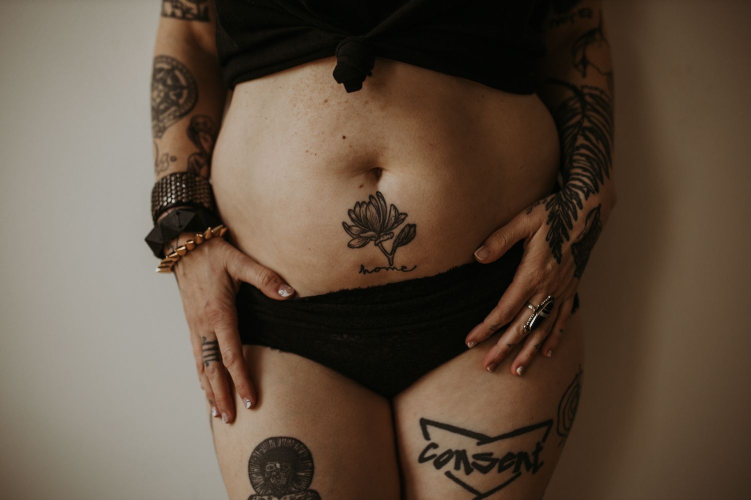 Close-up of a heavily tattooed woman's torso wearing black underwear, showcasing intricate black ink tattoo artwork.