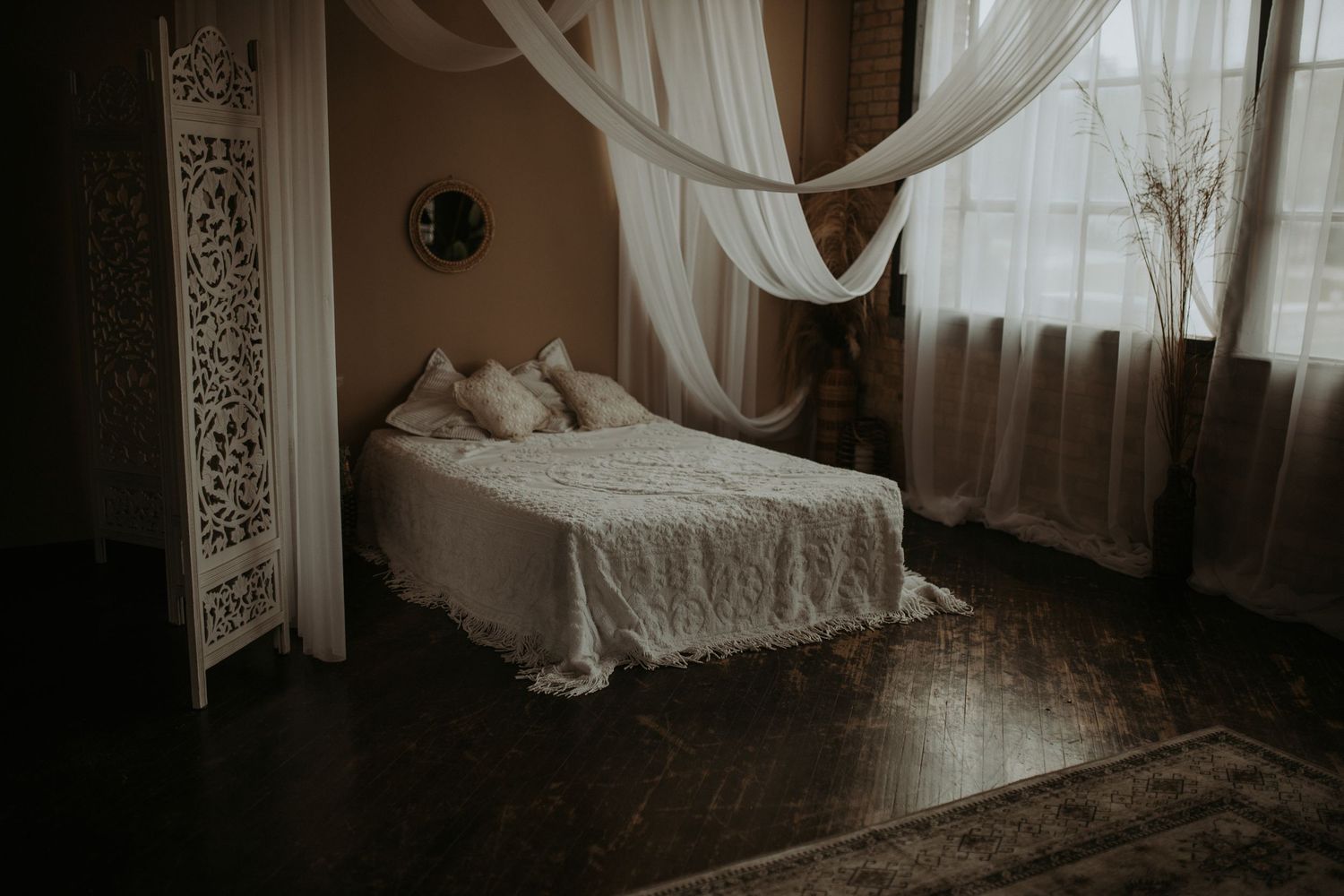 A romantic bedroom with white linens, sheer curtains, and dark hardwood floors in moody lighting.