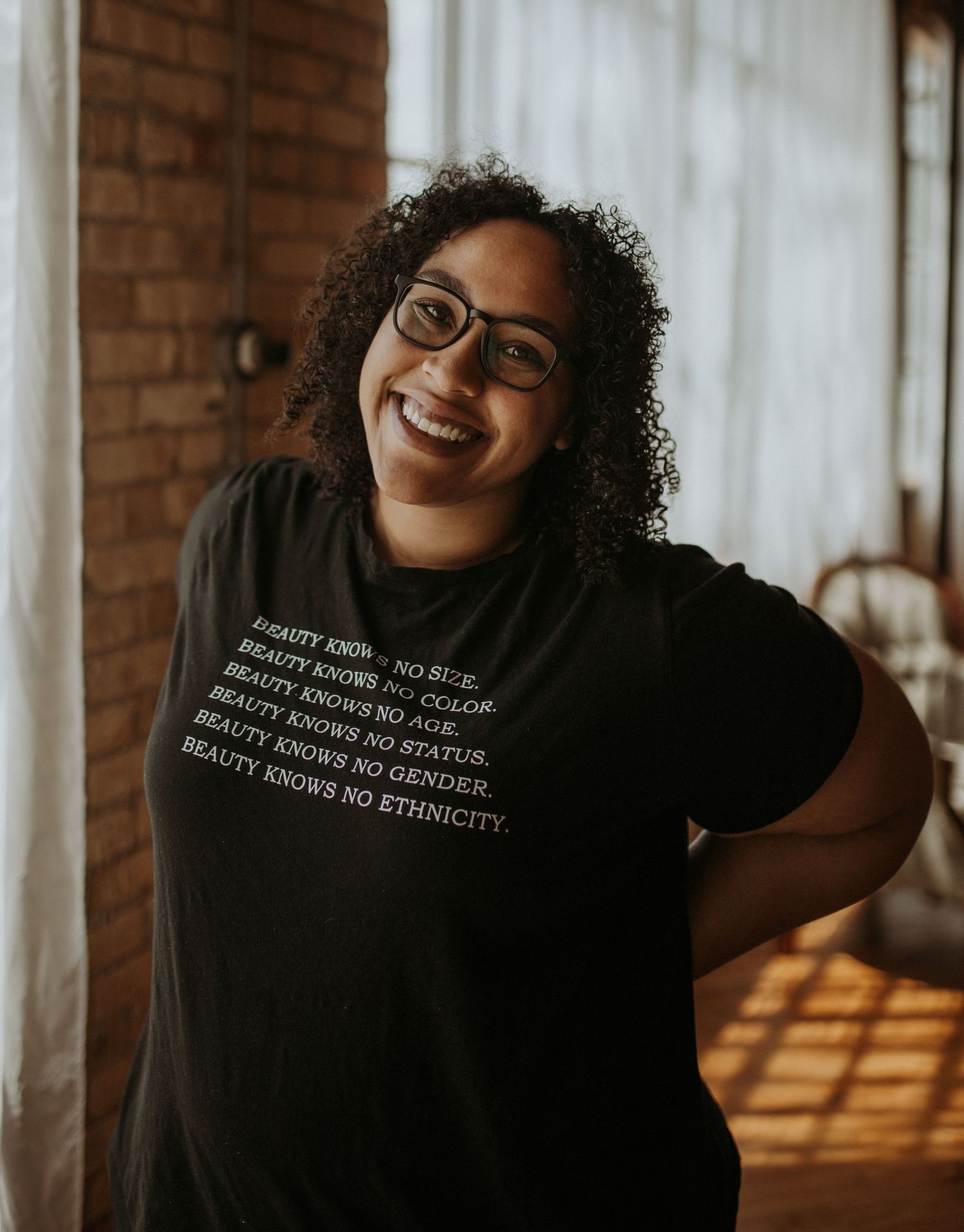 Smiling woman wearing black graphic tee reading Beauty Knows No Size Color Age Status Gender Ethnicity posing indoors.