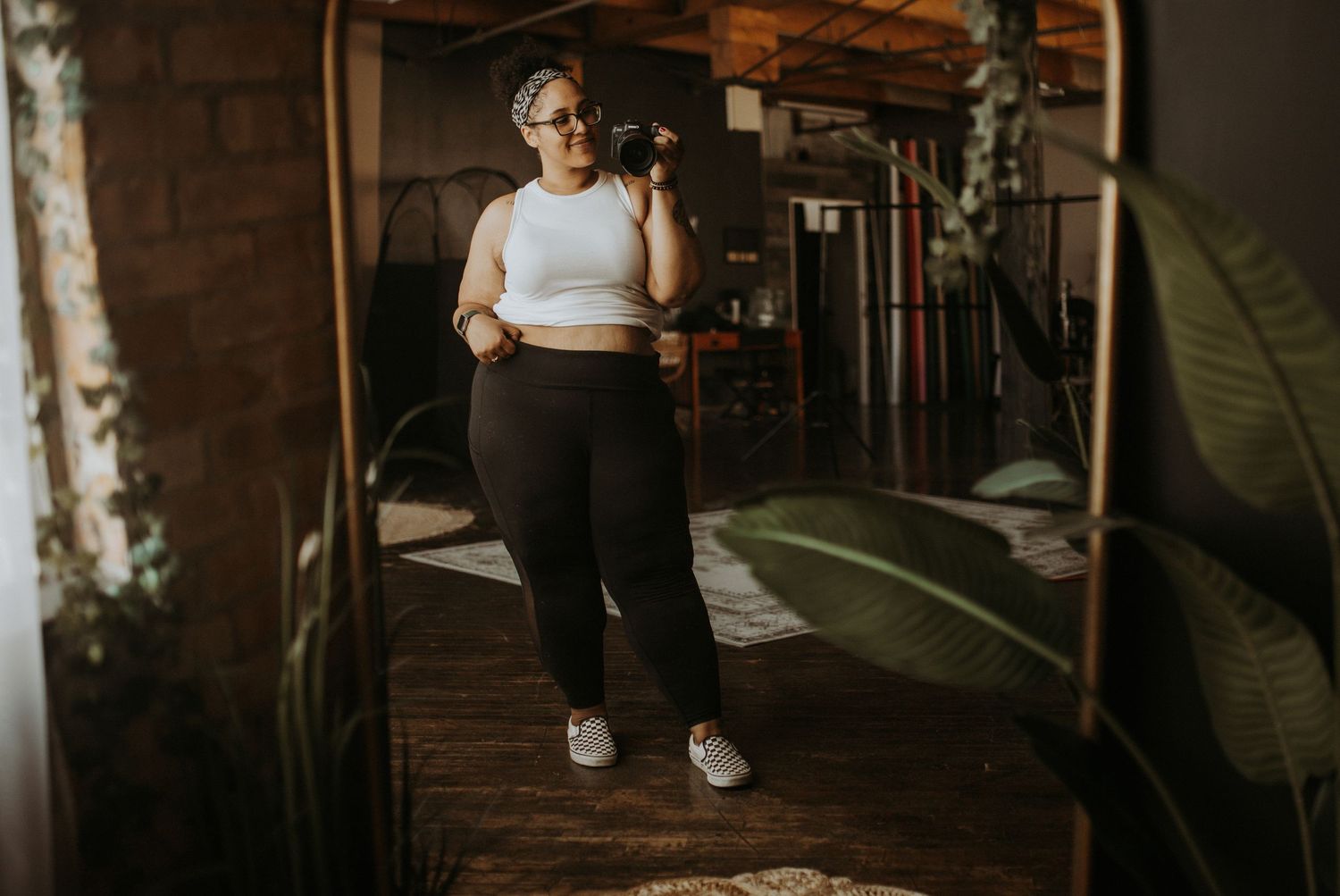 A plus-size woman in white crop top and black leggings holds a camera while standing in a stylish loft space.