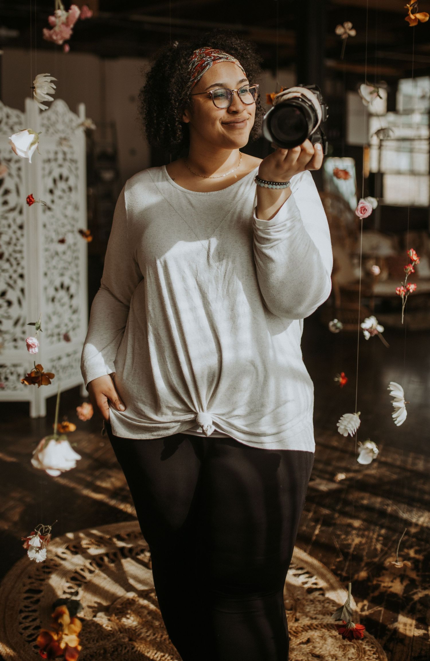 Photographer wearing glasses and white sweater holds camera while smiling in moody indoor setting with fairy lights.