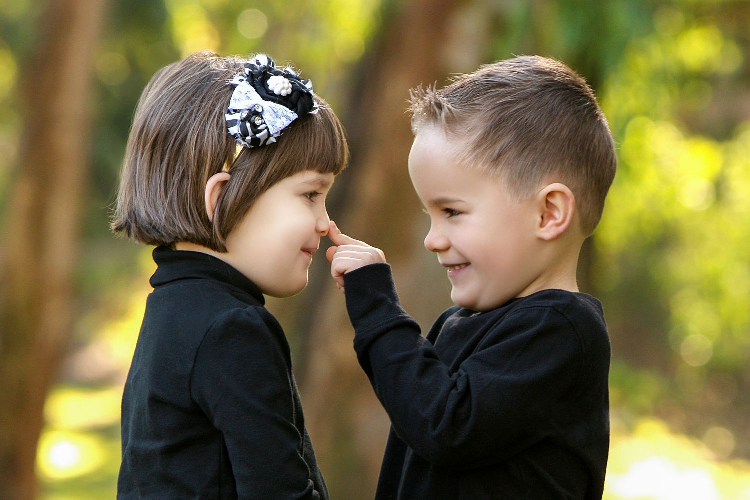 Two young children in black outfits share a playful moment outdoors, with a boy gently touching a girl's nose.