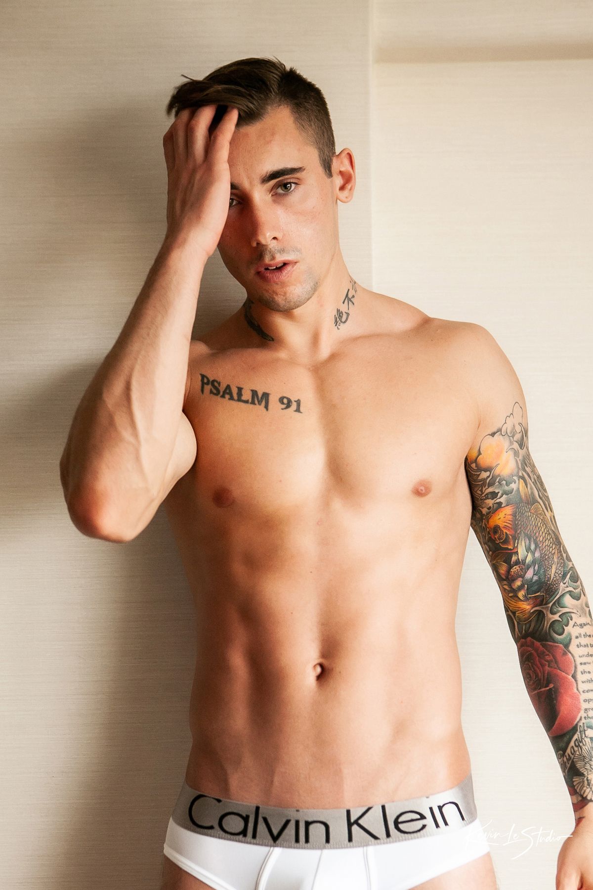 Shirtless model wearing Calvin Klein underwear poses against beige wall showing tattoos and toned physique.