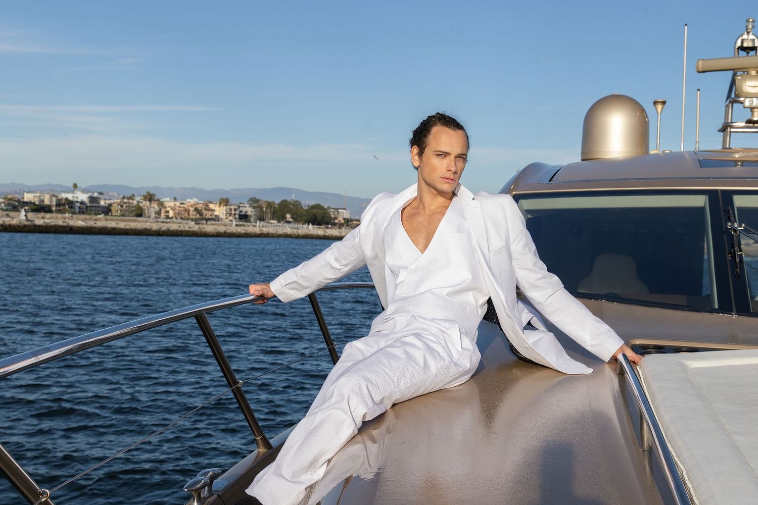 Person in white shirt sailing a boat on calm blue waters near a coastal shoreline.