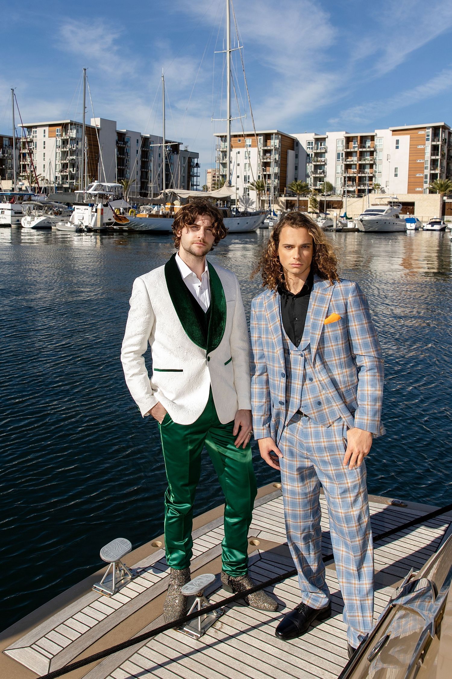 Two people in stylish suits pose on a dock at a marina with luxury apartments and boats in the background.