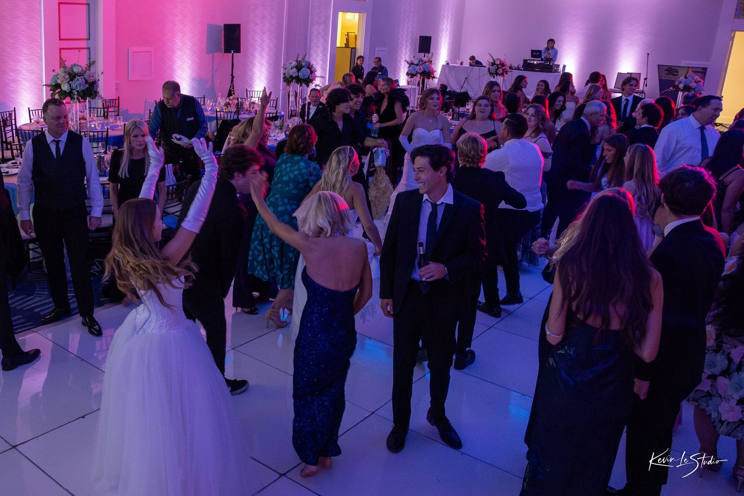 Wedding reception guests dancing on a white dance floor illuminated by purple lighting at an elegant venue.