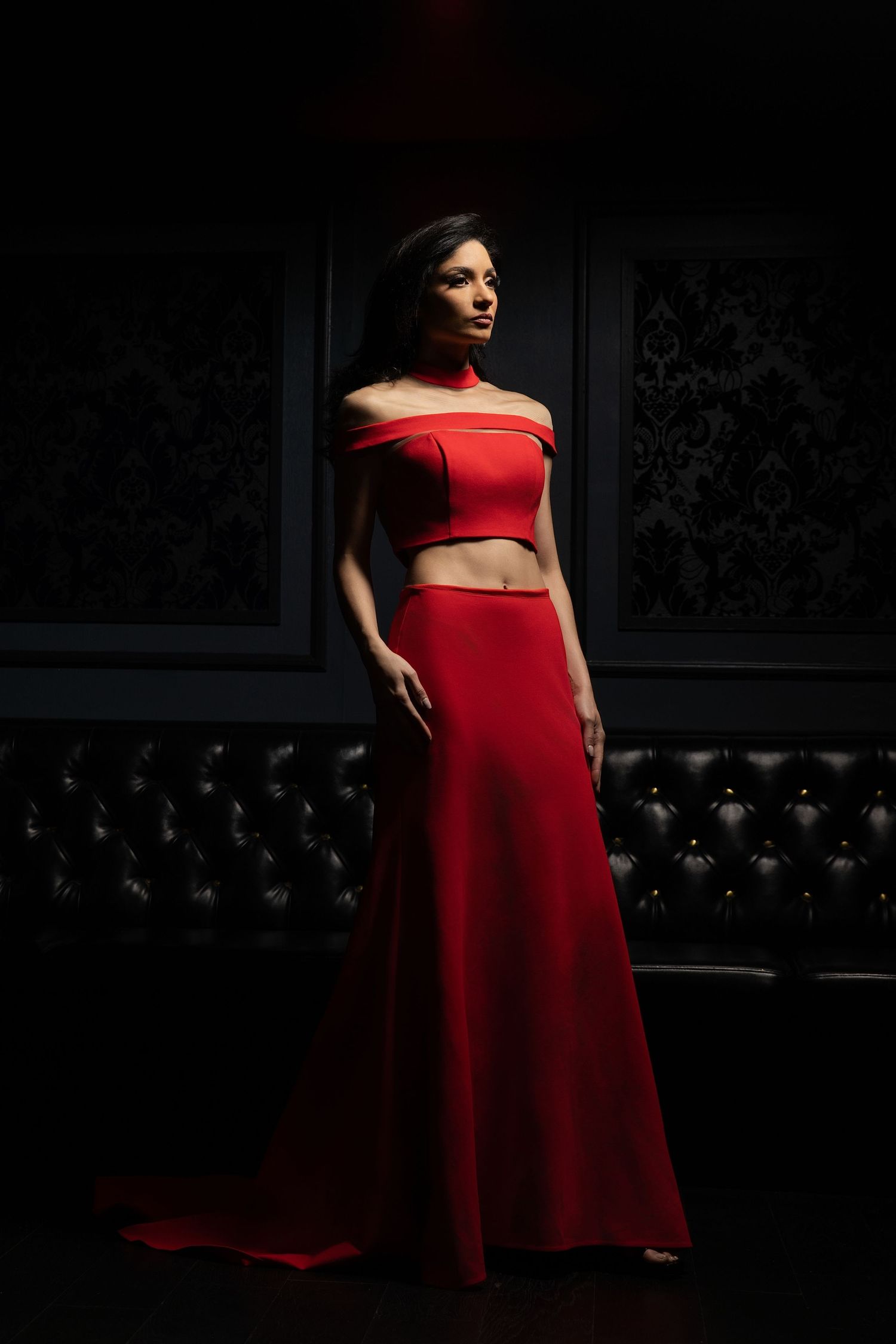 A model in a striking red two-piece evening gown poses dramatically against a dark background with tufted upholstery.