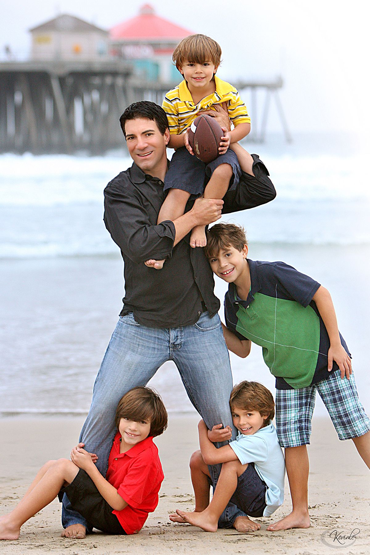 A father poses on a beach with four young sons, one held overhead with a football, others gathered around him playfully.