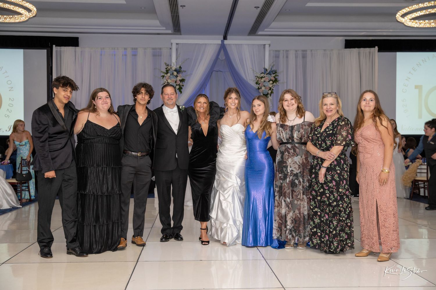 A group of formally dressed people pose together at an elegant event in a ballroom with decorative lighting.