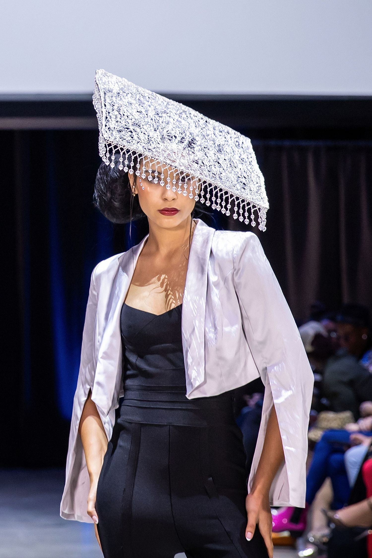 Fashion model walks runway in black fitted dress with white satin cape and dramatic beaded headpiece at fashion show.