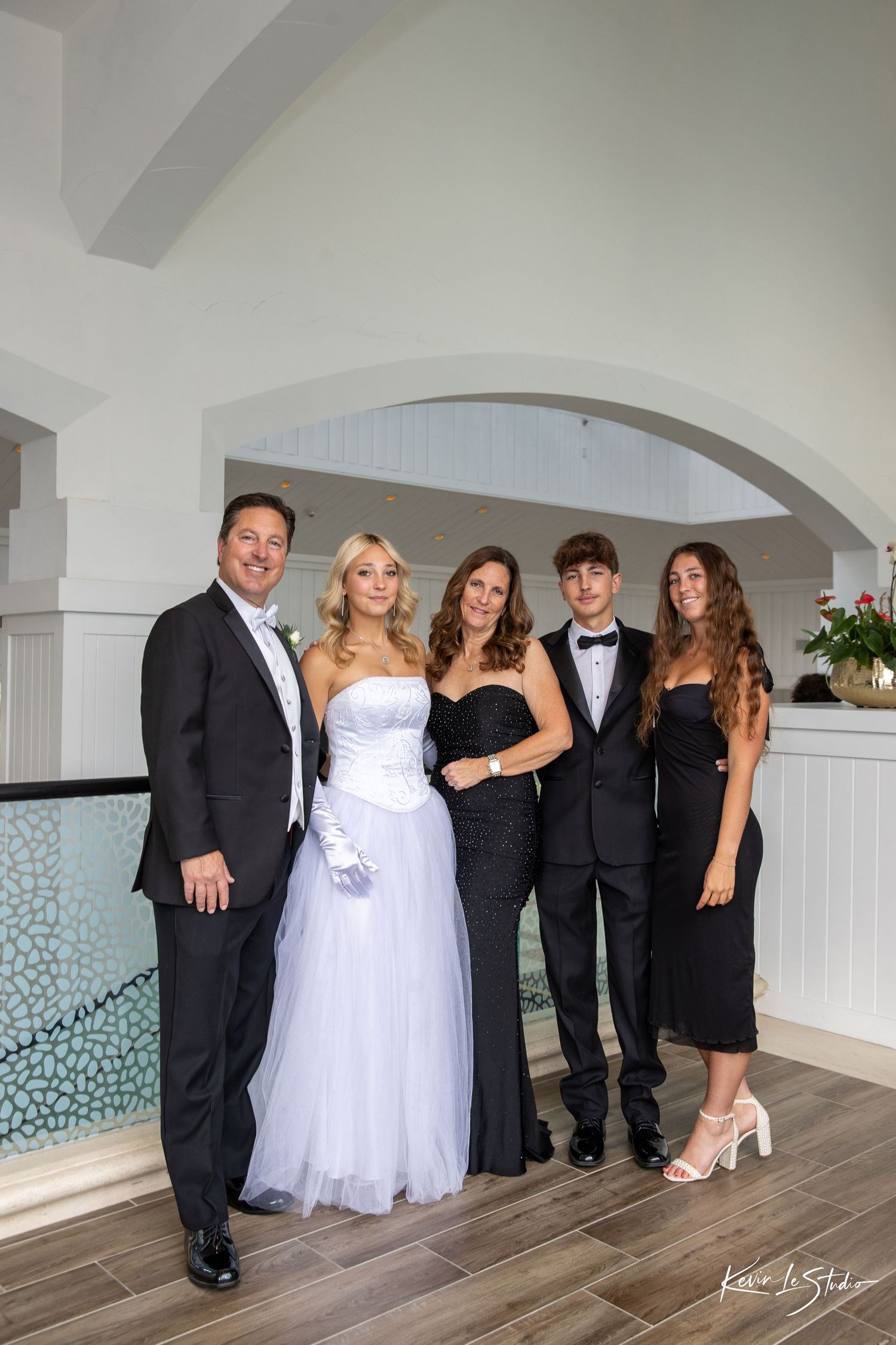 Wedding party poses together in formal attire along a white interior wall with decorative mosaic tiles.
