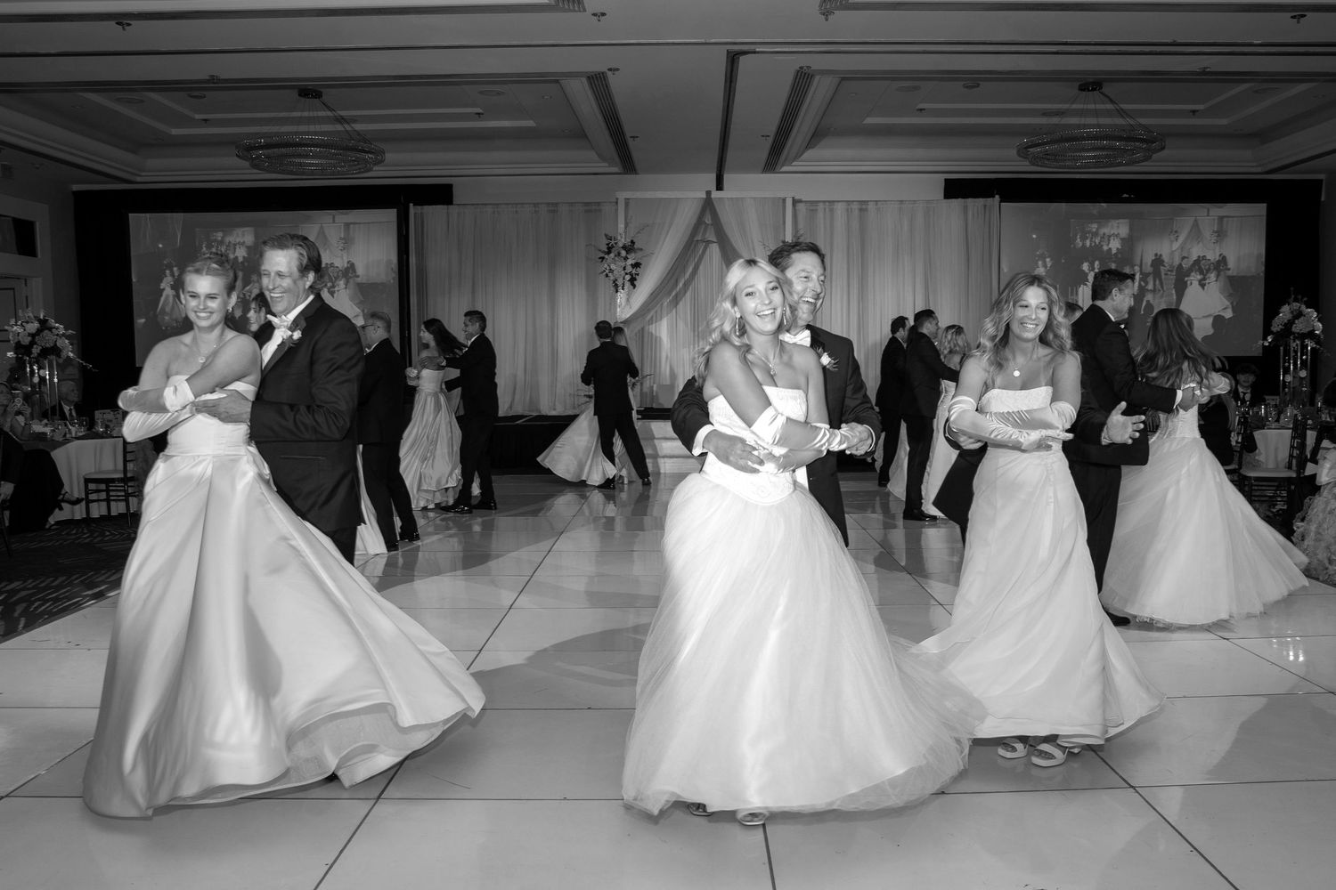 Black and white photo of couples dancing in formal attire at an elegant ballroom event.