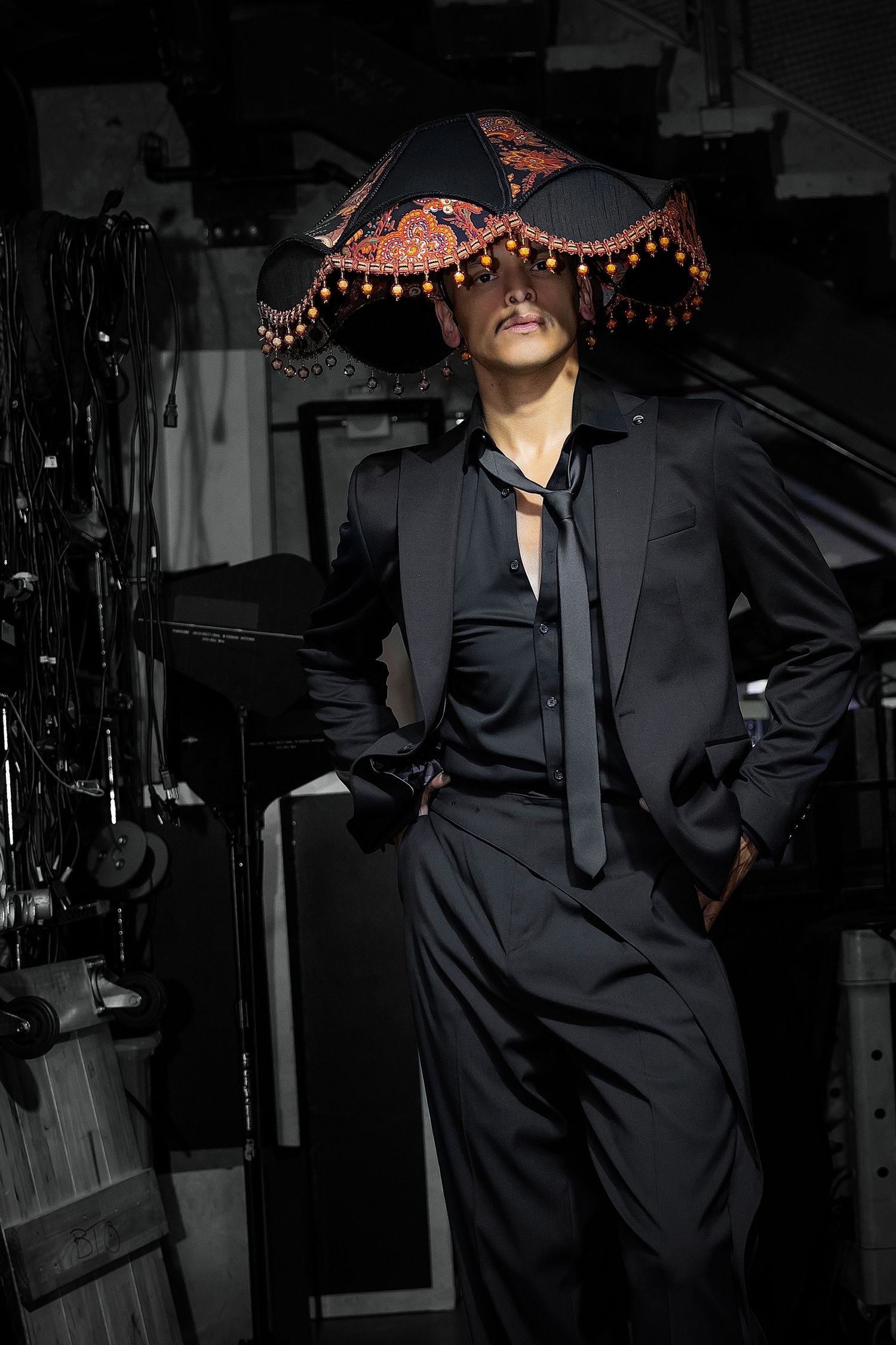 A dramatic fashion photo showing a black suit and ornate beaded hat with pearls in a dark moody setting.