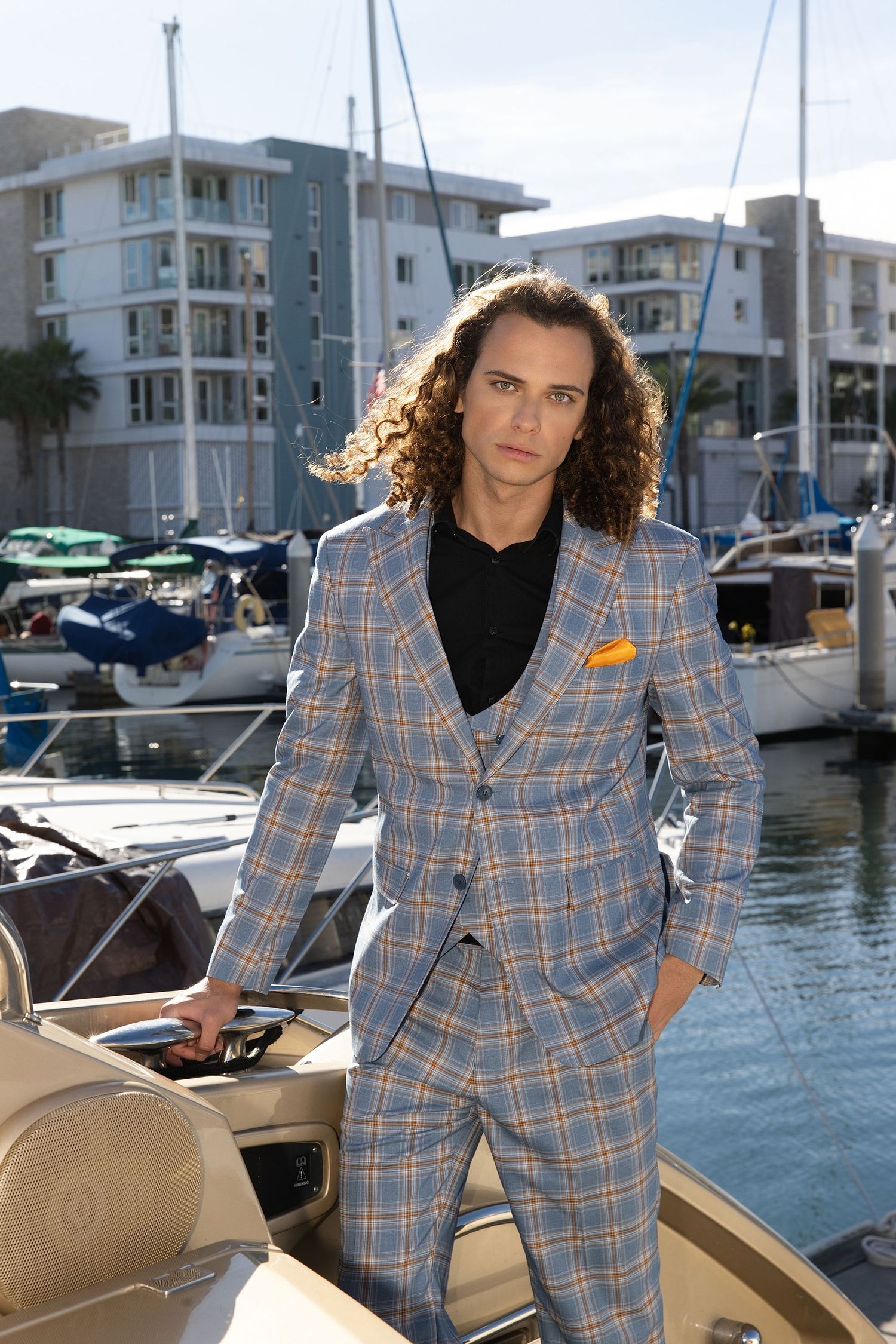A person in a plaid blazer suit poses on a boat dock with waterfront condominiums and marina in the background.