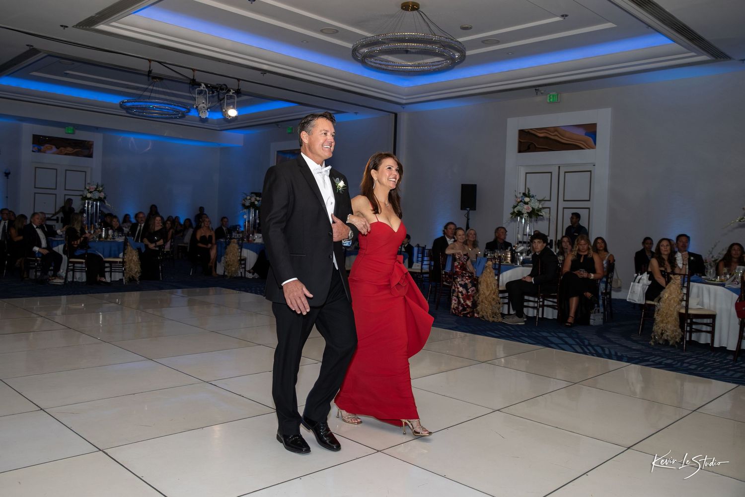 Dancers perform a series of elegant dance moves on a white floor under blue lighting at a formal event.