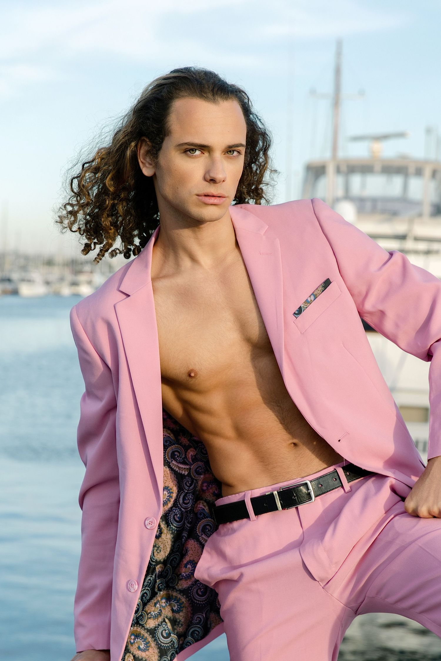 Model wearing pink blazer with shirtless styling poses against marina backdrop with boats and water.