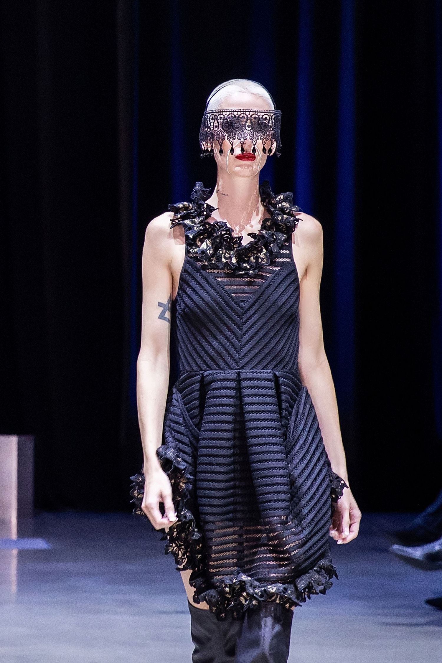 Fashion model on runway wearing black striped cocktail dress with floral embellishments and metallic headpiece.