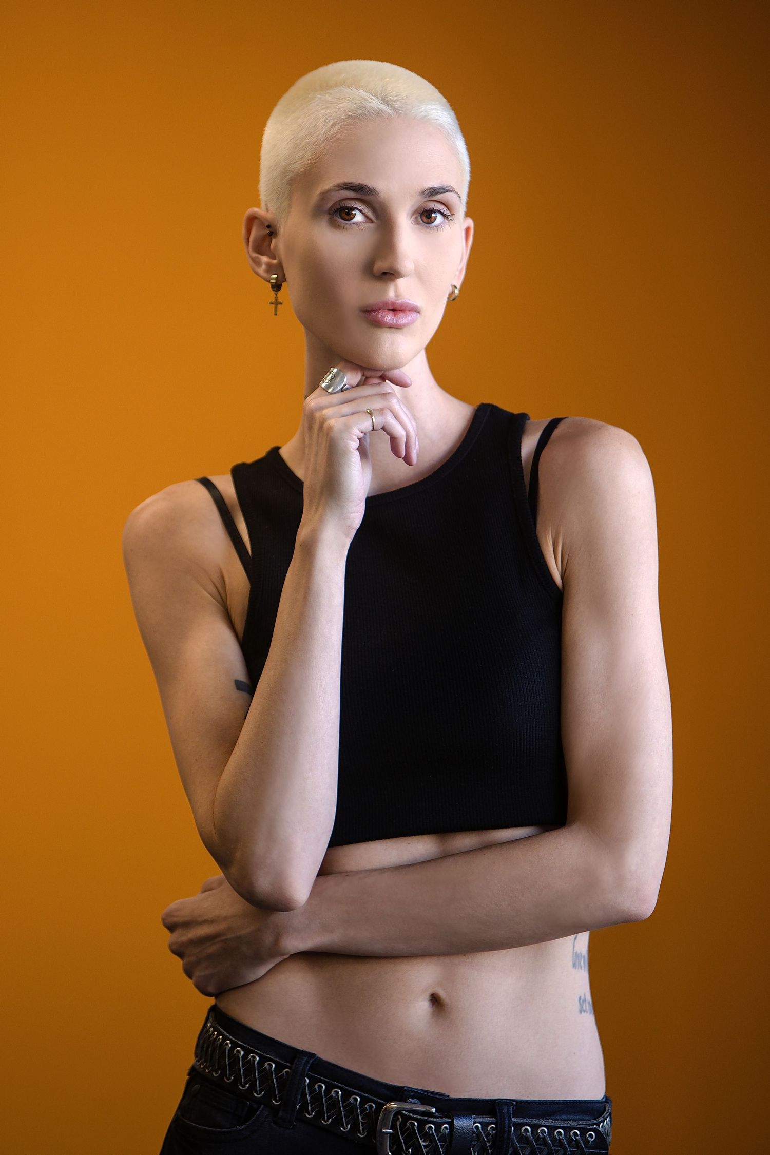 Studio portrait against orange background showing person with platinum blonde hair in black crop top in thoughtful pose.