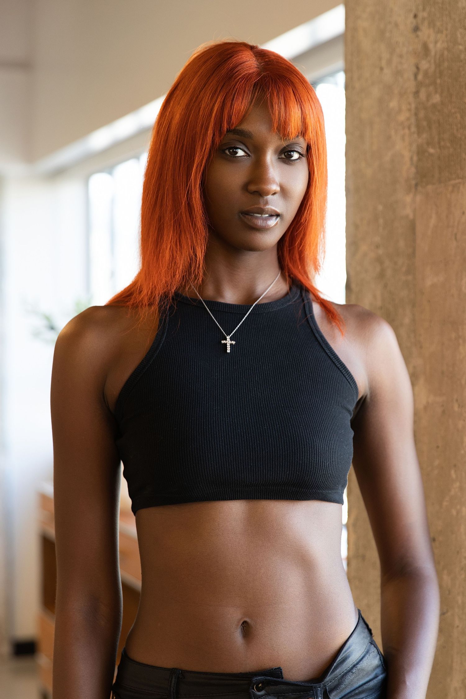 Athletic person with bright orange hair wearing black workout attire poses in a sunlit industrial space.