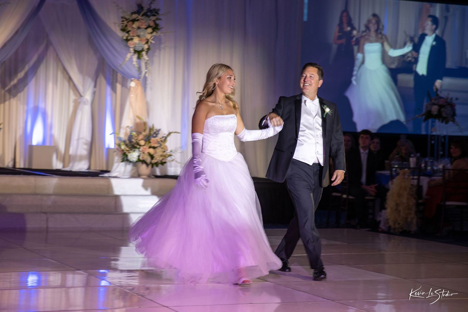 A couple performs an elegant dance in formal attire on an illuminated ballroom floor during their wedding reception.