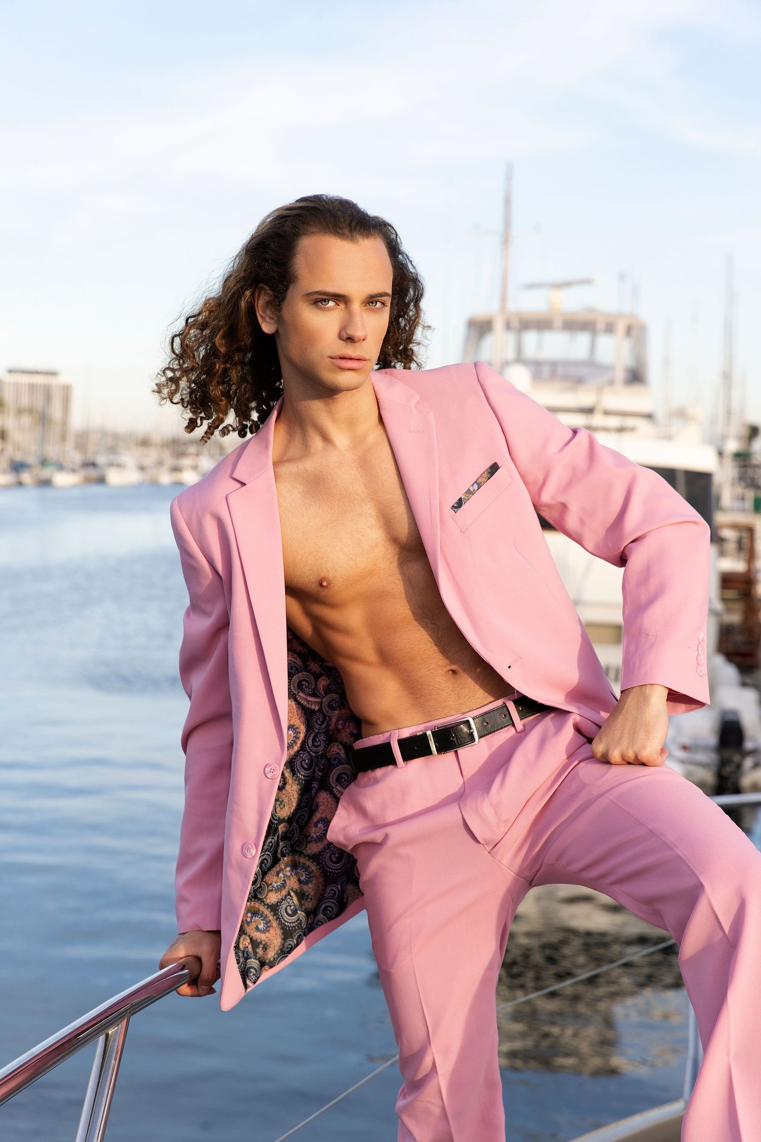 Man in open pink suit jacket and pants poses on yacht dock with marina background.