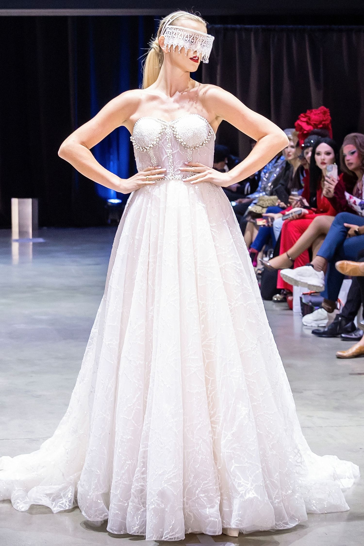 White lace wedding gown with full skirt shown from multiple angles on fashion runway with audience in background.