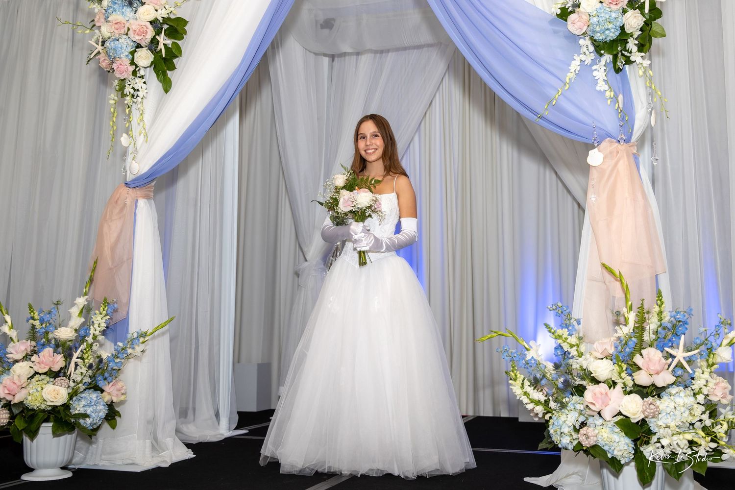 A white wedding dress with tulle skirt displayed against an elegant backdrop of white drapes and floral arrangements.