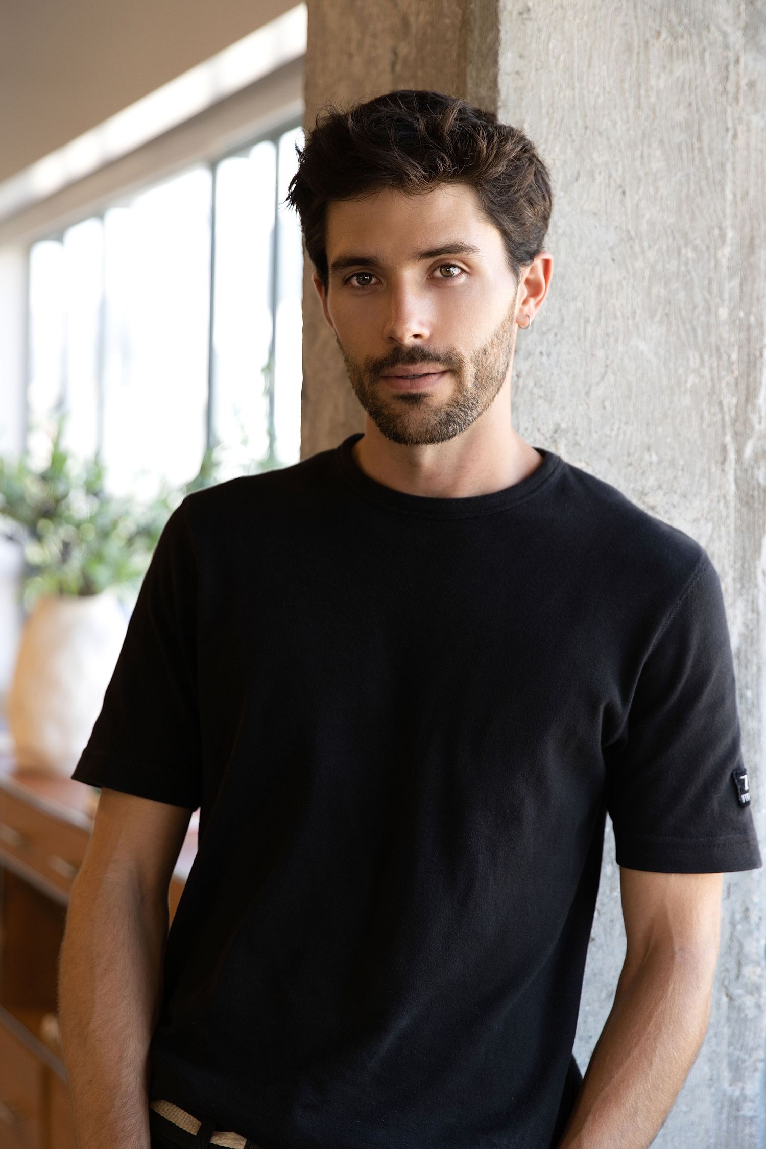 A person in a black t-shirt smiles while sitting against a concrete wall with natural lighting streaming in.