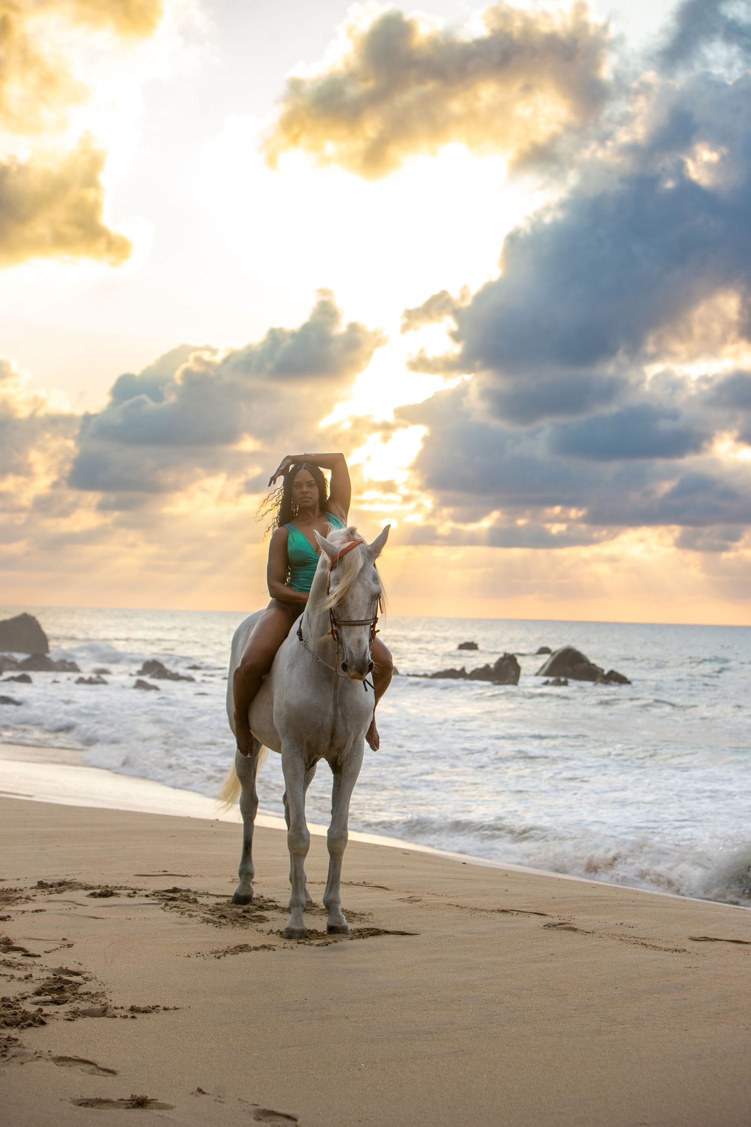 A white horse stands on a sandy beach at sunset with crashing waves and dramatic cloudy sky in the background.