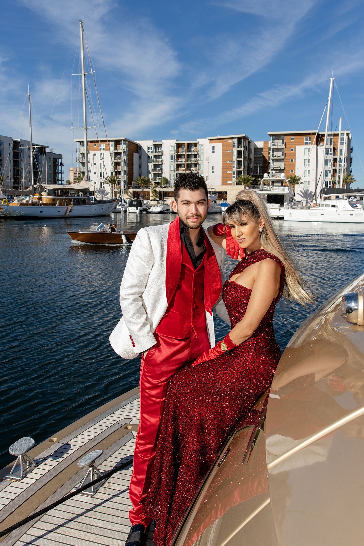 A couple in coordinated red and white outfits pose together on a yacht at a marina with apartment buildings in background.