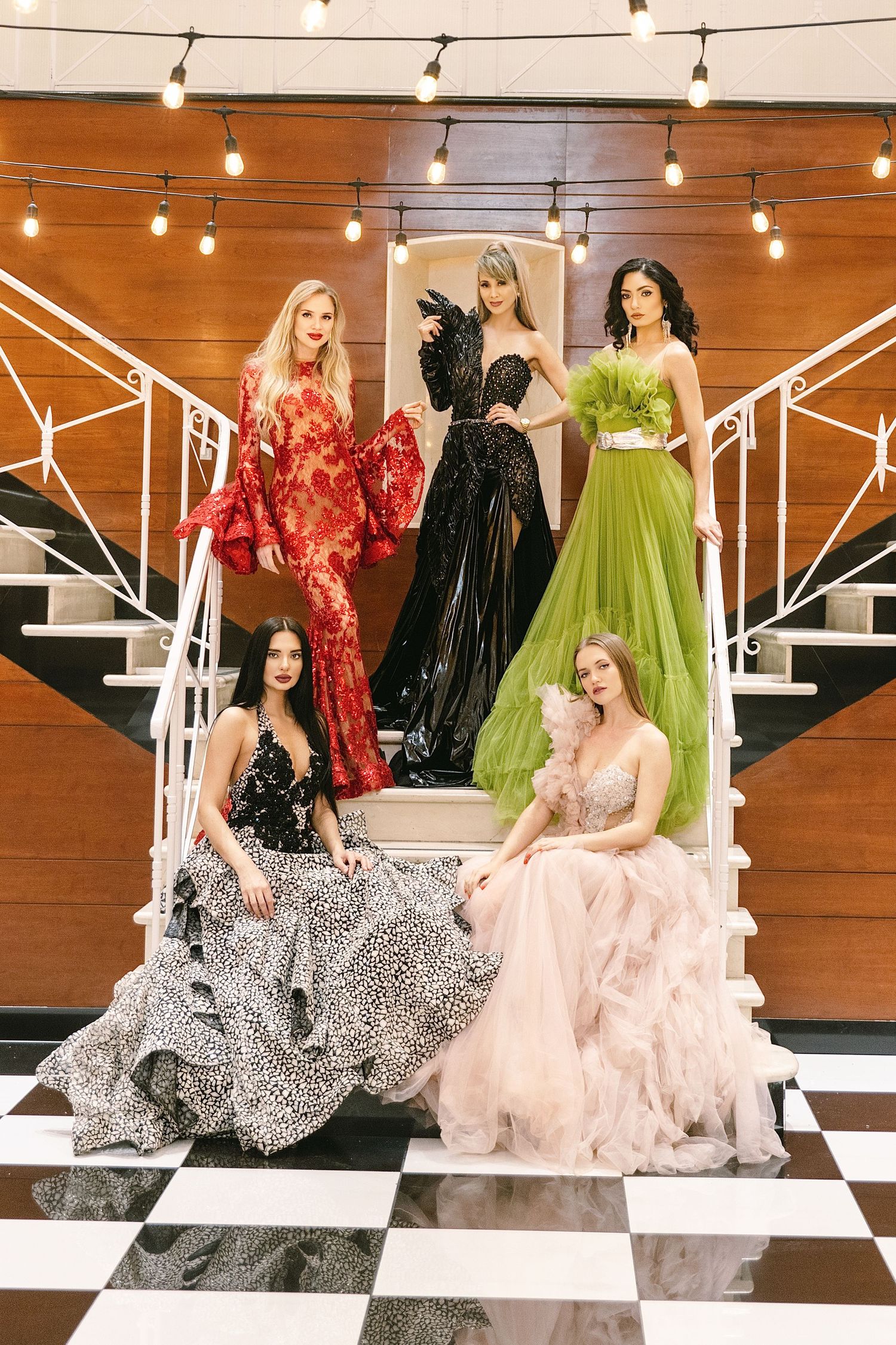 Models showcase glamorous evening gowns in red, black, green, and pink against a rustic wooden backdrop with string lights.