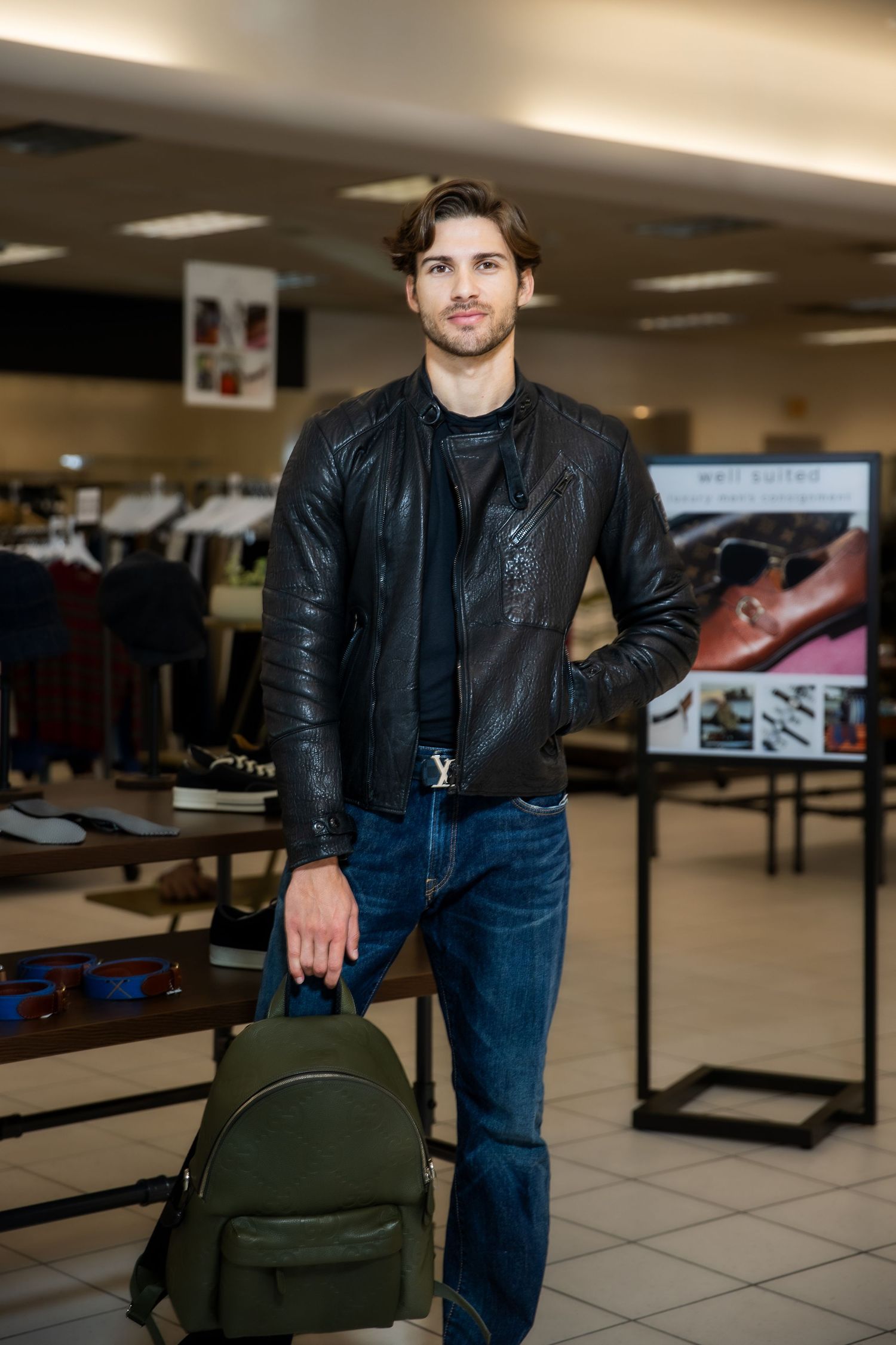 A person wearing a black leather jacket and jeans stands in an indoor space holding a dark green backpack.