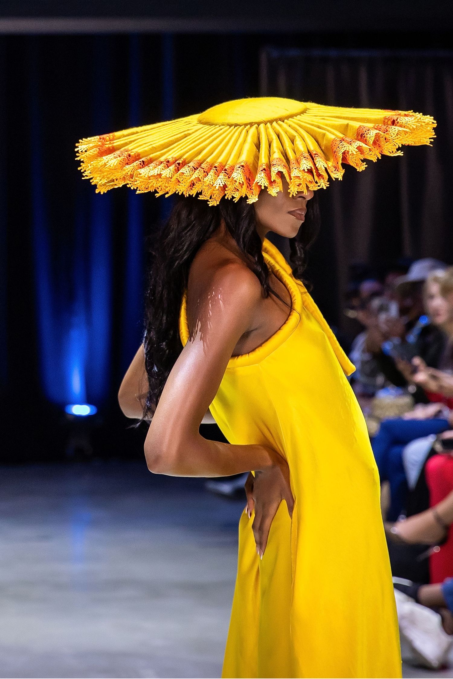 Fashion model on runway wearing bright yellow dress and dramatic matching fan-shaped hat during fashion show.