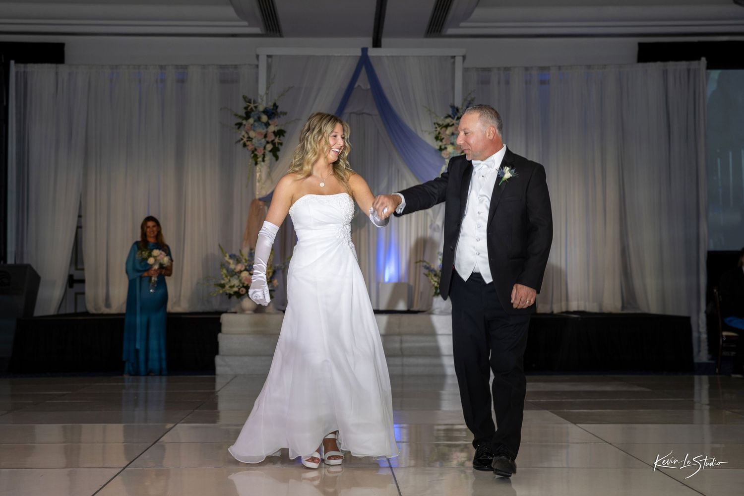 A couple shares a dance moment at their wedding reception with a person in a teal dress visible in the background.