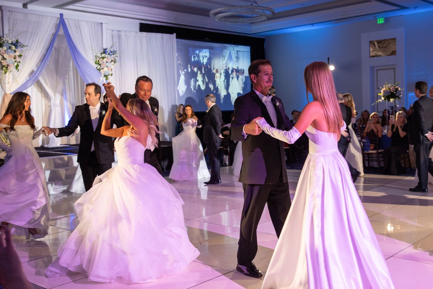 Elegant couples dancing at a wedding reception with white decor and blue lighting creating a romantic atmosphere.