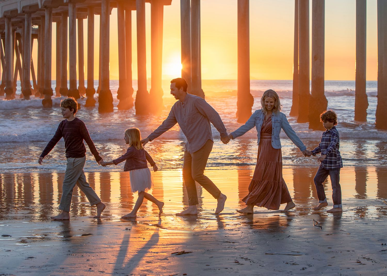 Family of five holding hands walking on beach at golden sunset near pier with ocean waves.