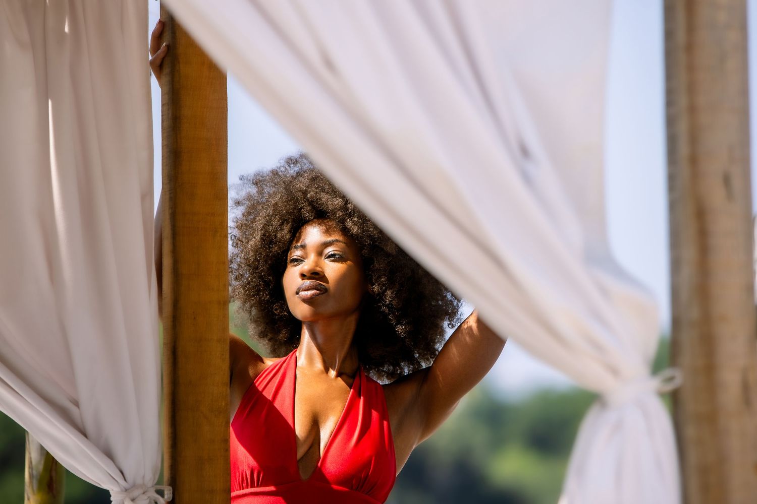 Woman in red dress poses gracefully against white curtains in a natural light setting.