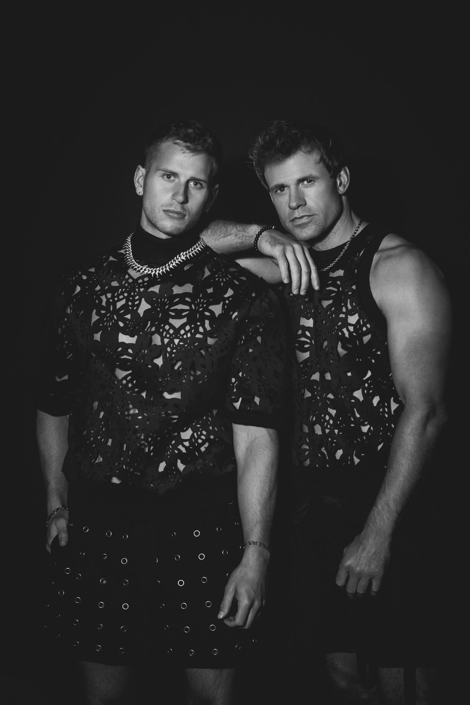 Two people pose together wearing matching black t-shirts with geometric patterns in a dramatic black and white photo.
