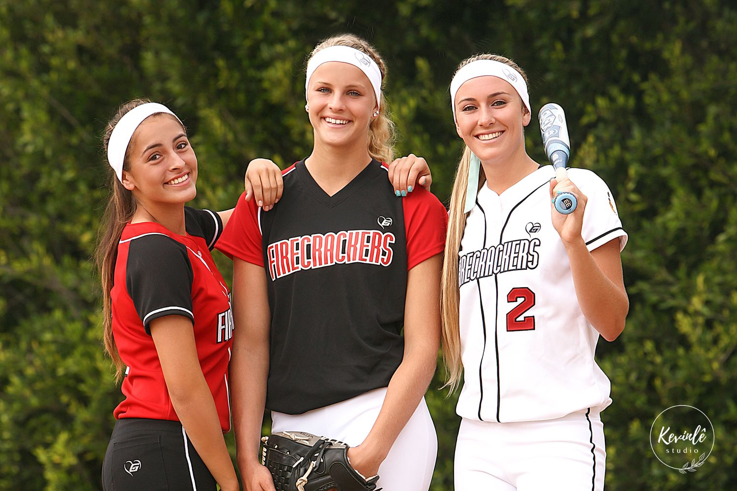A group of softball players in red and white uniforms pose together on a sunny day by trees.