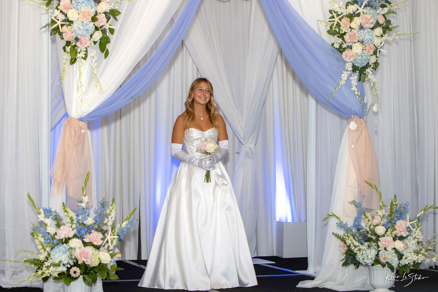 A wedding ceremony setup with white draping, floral arrangements, and elegant decor against a light backdrop.