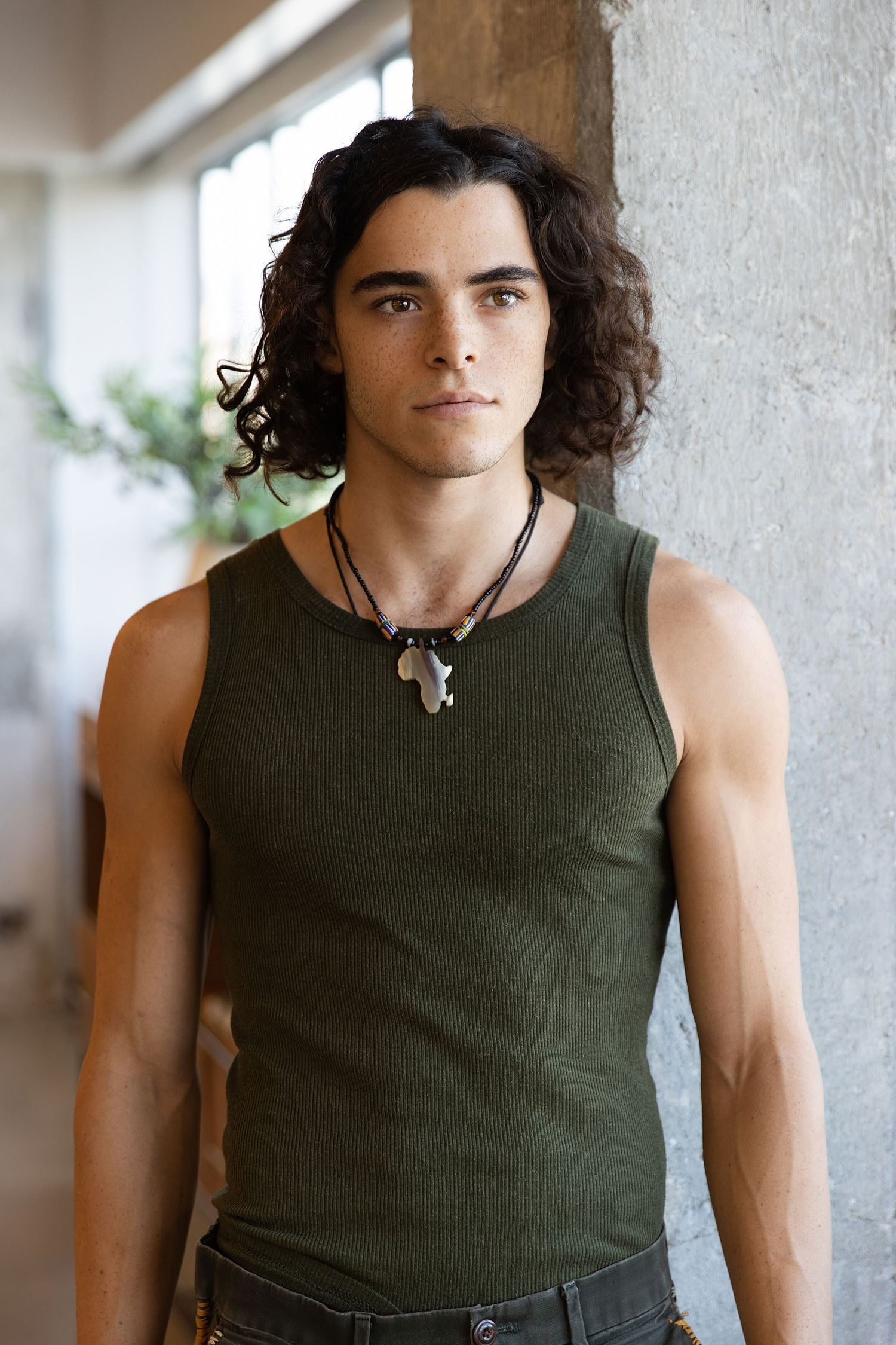 Young person with long dark curly hair wearing an olive green tank top and necklace leans against wall in natural light.