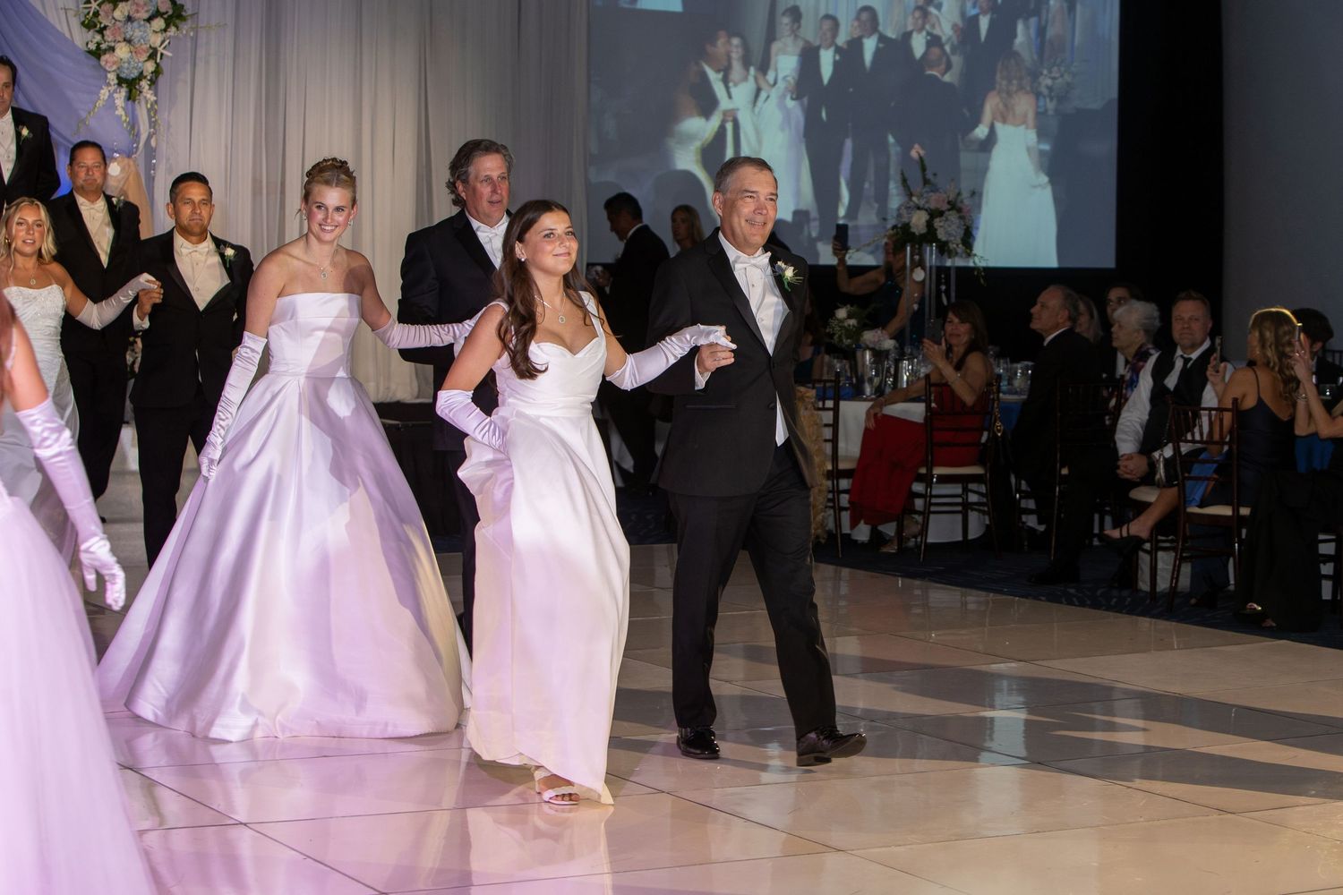Dancers in formal attire perform at an elegant ballroom event with white and lavender dresses flowing gracefully.