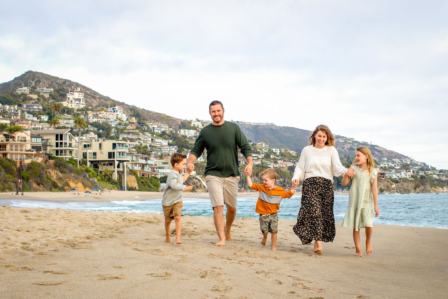 Family walking along scenic coastal path with mountains and beach homes in background during sunset.