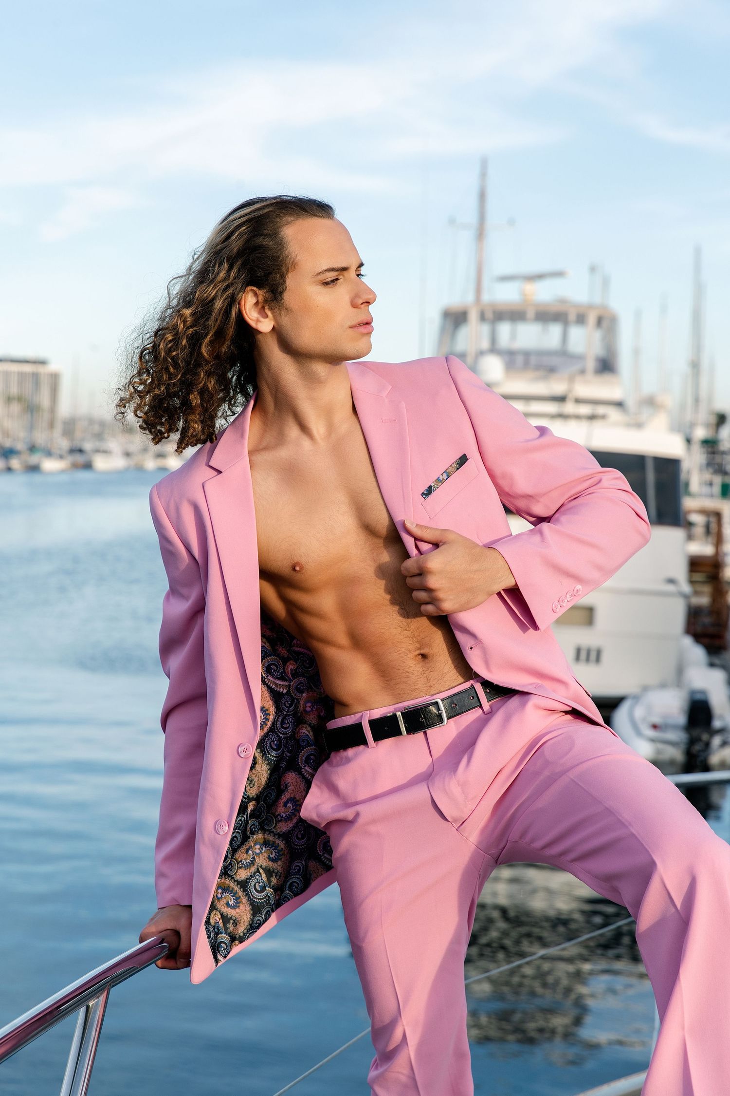 Model in pink suit poses shirtless on yacht deck with marina and boats in background.