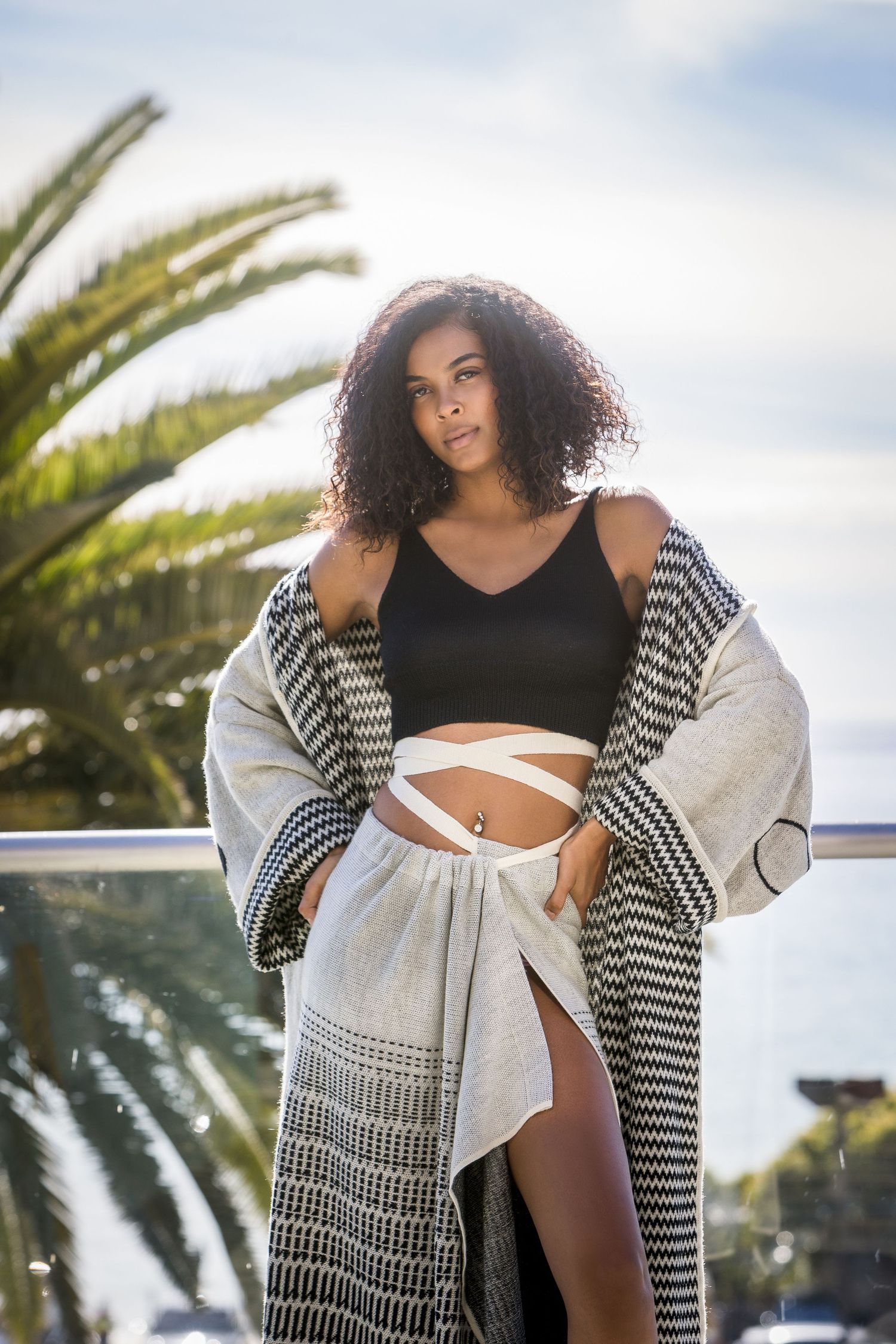 A model poses outdoors in a black and white striped cardigan and swimwear against a palm tree backdrop.