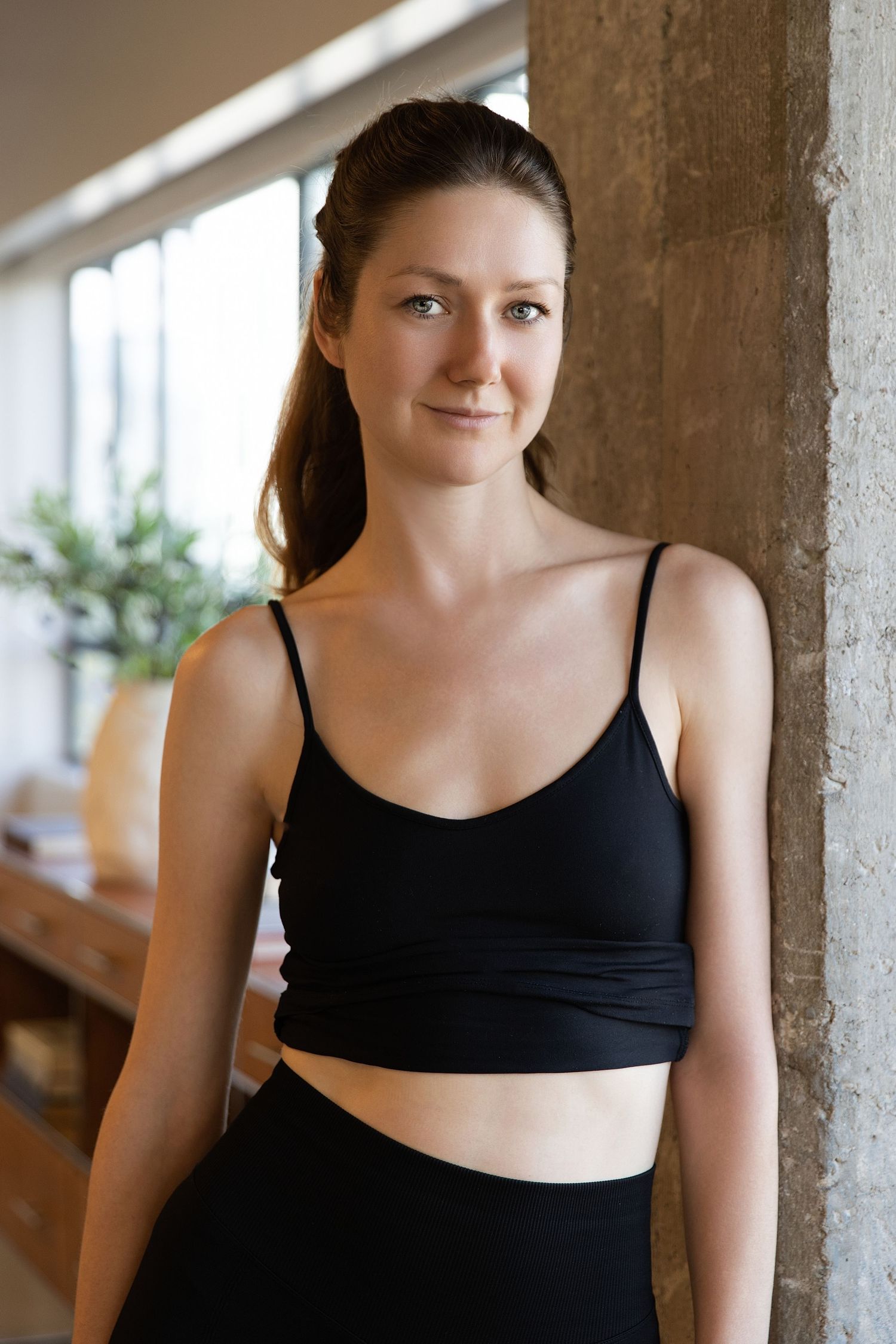 Woman in black tank top posing against concrete wall in natural lighting.