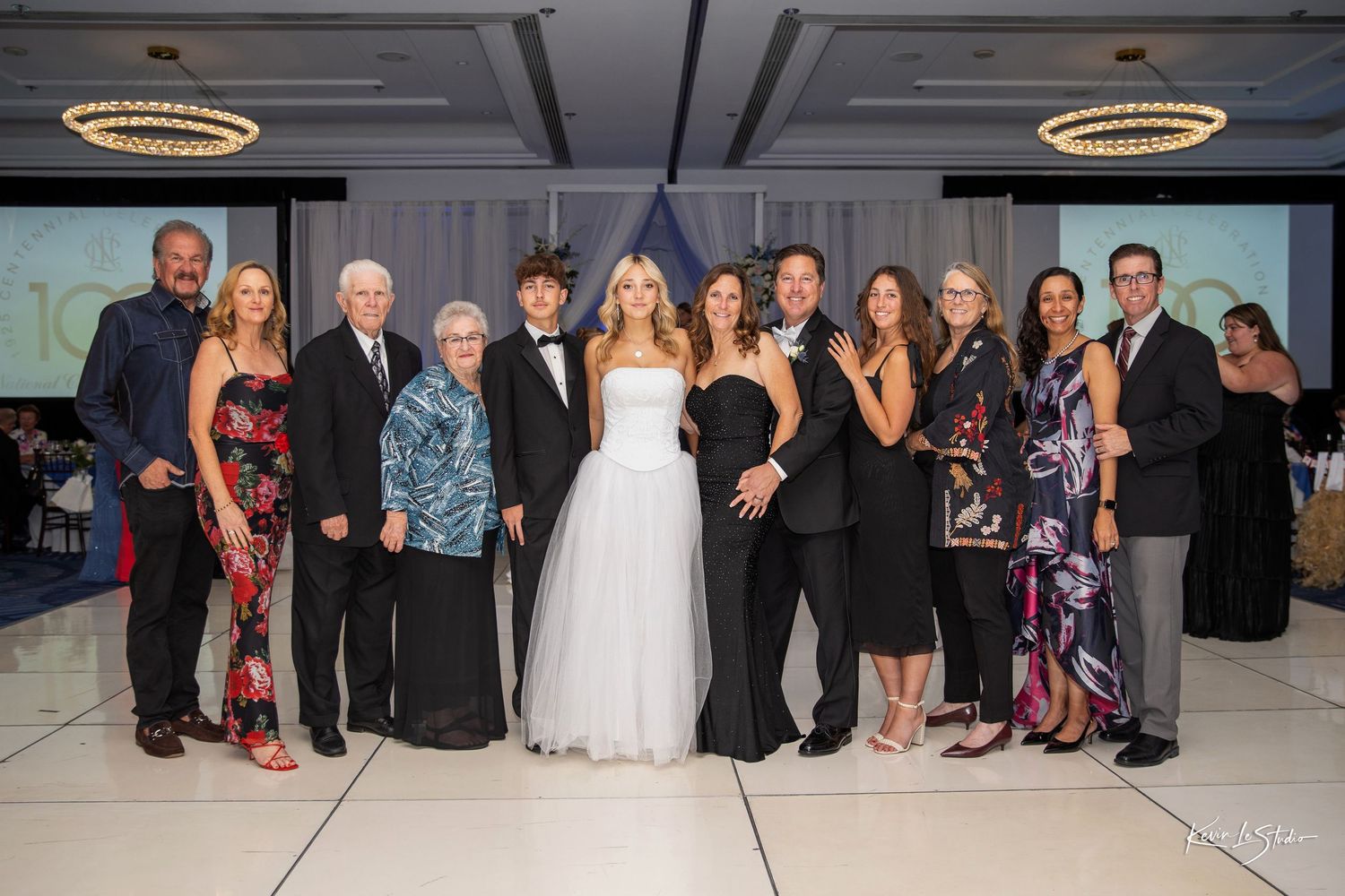 A group of wedding guests pose together on a dance floor at a formal reception celebration.