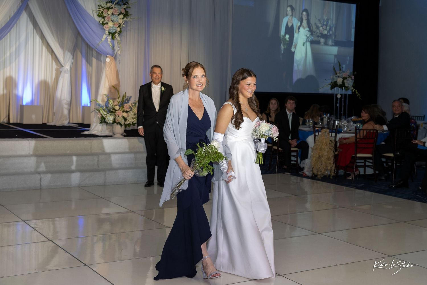 Wedding party members walk across a polished dance floor during a reception with decorative white draping and projector screen.