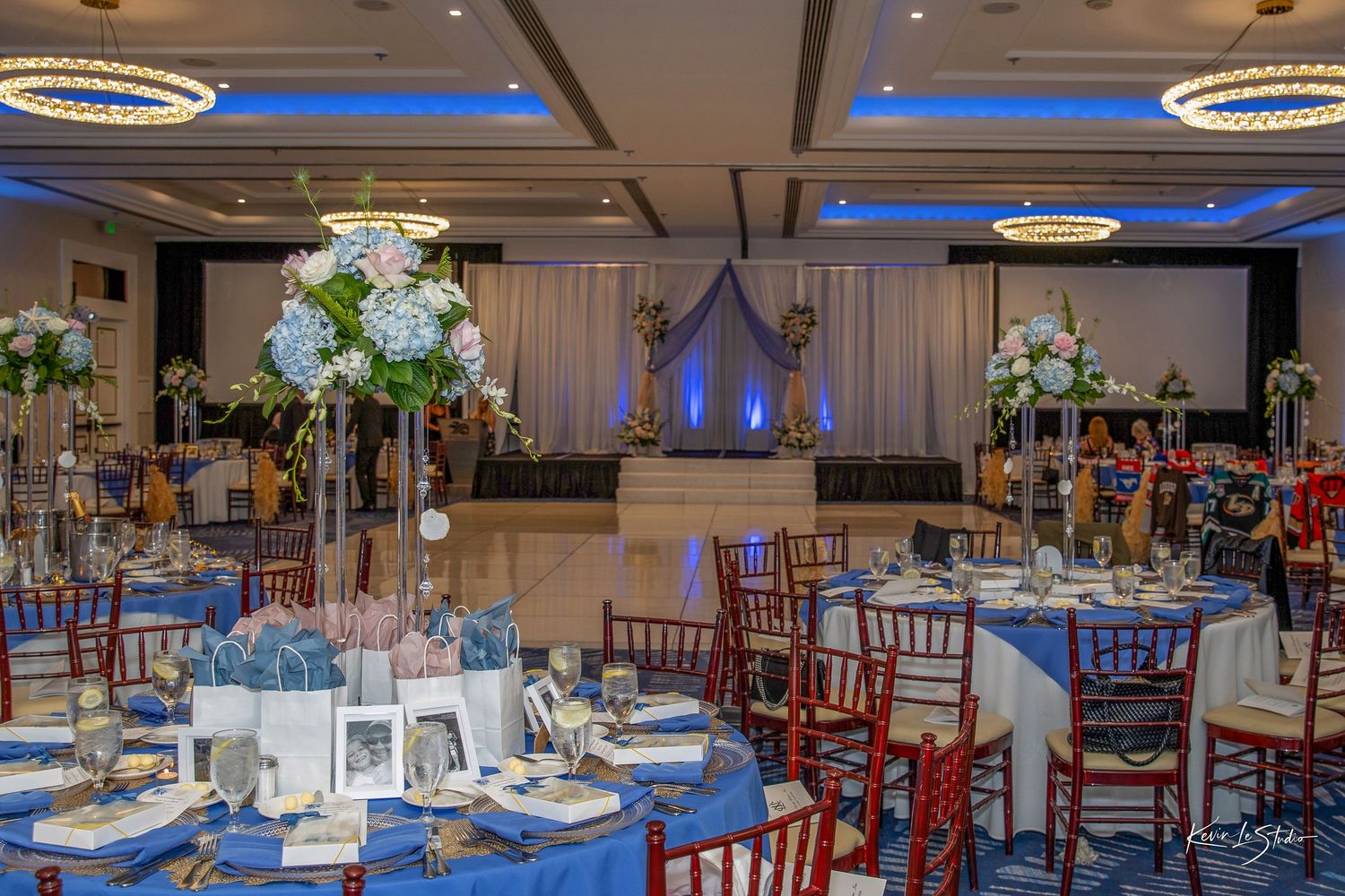 Elegant wedding reception ballroom with blue uplighting, tall floral centerpieces, and mahogany chiavari chairs.